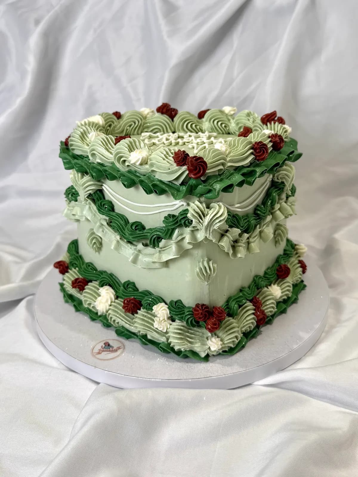 A heart-shaped vintage cake piped in sage green with red cherries and scalloped borders