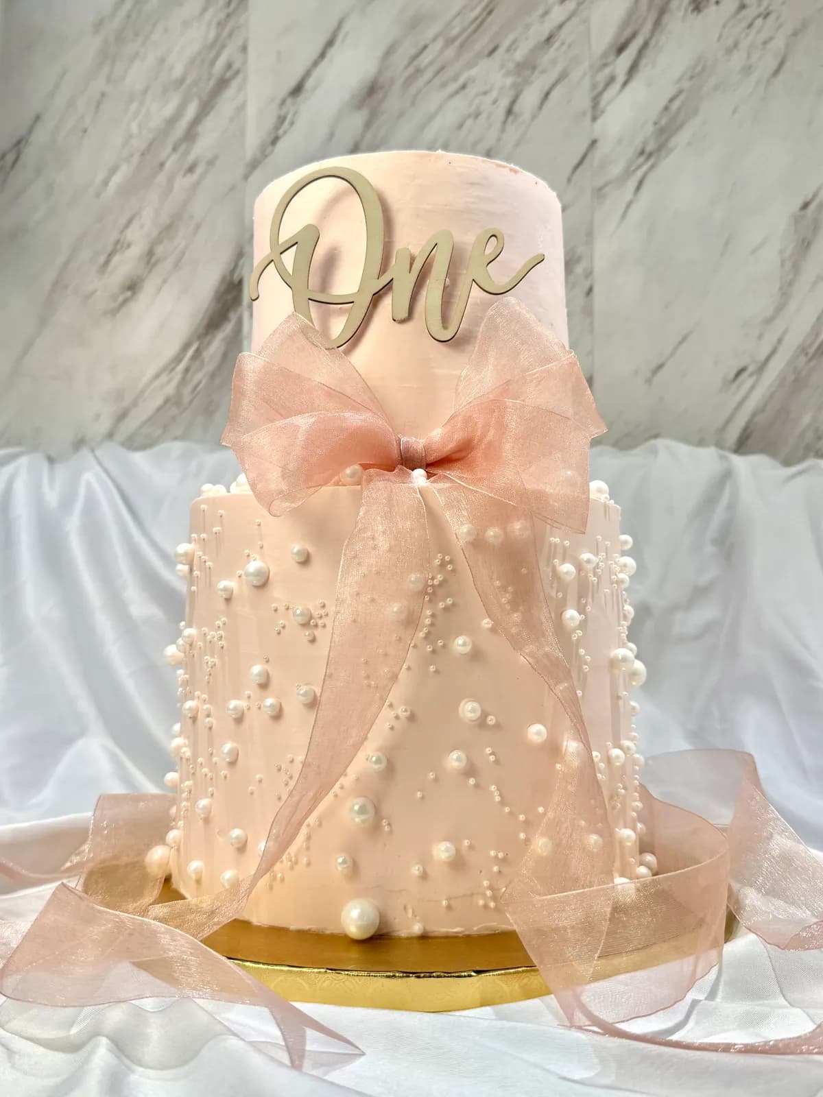 A blush first-birthday cake wrapped in organza with a satin bow and rows of pearls